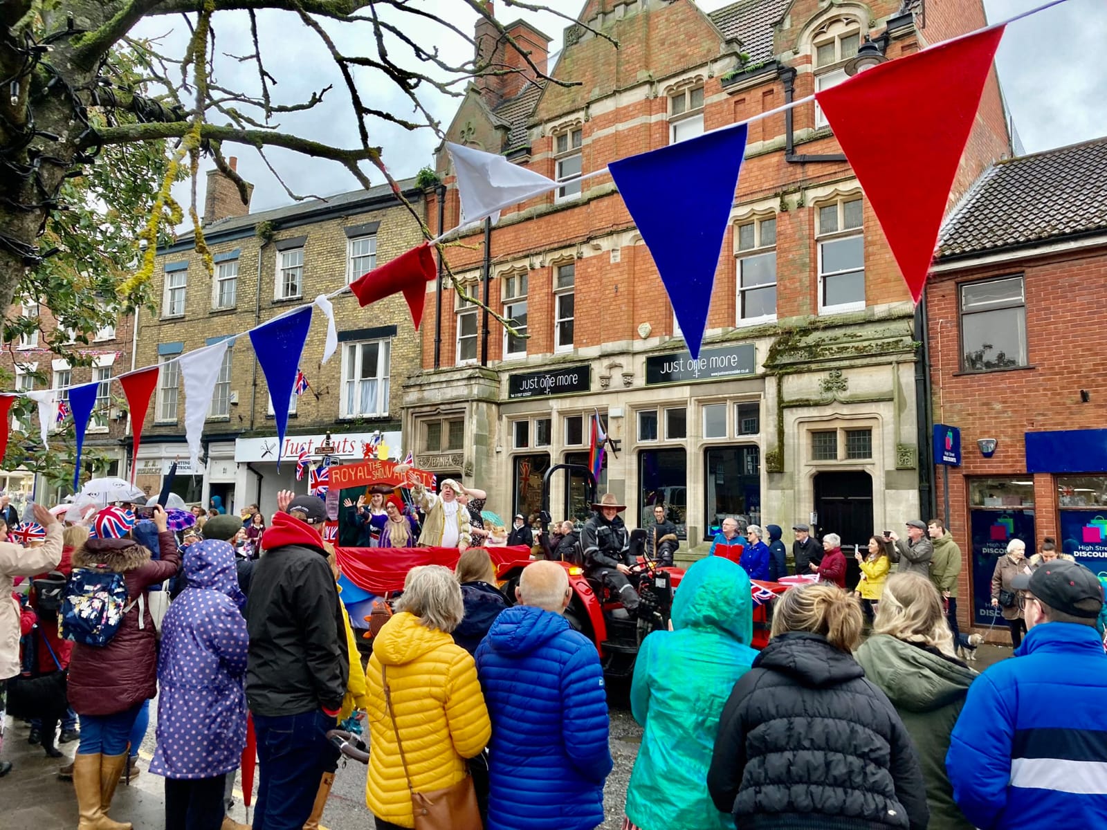 image of a parade down the High Street in front of Just One More Bike. People dressed in colourful waterproof clothing in the foreground. The parade float that is seen in the image is from Horncastle Theatre Company, being pulled by a red tractor.